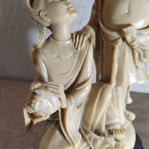May include: A white ceramic sculpture of two figures, a man and a woman, standing on a black base. The woman is kneeling and holding a bouquet of flowers. The man is standing behind her with his hand on her shoulder.