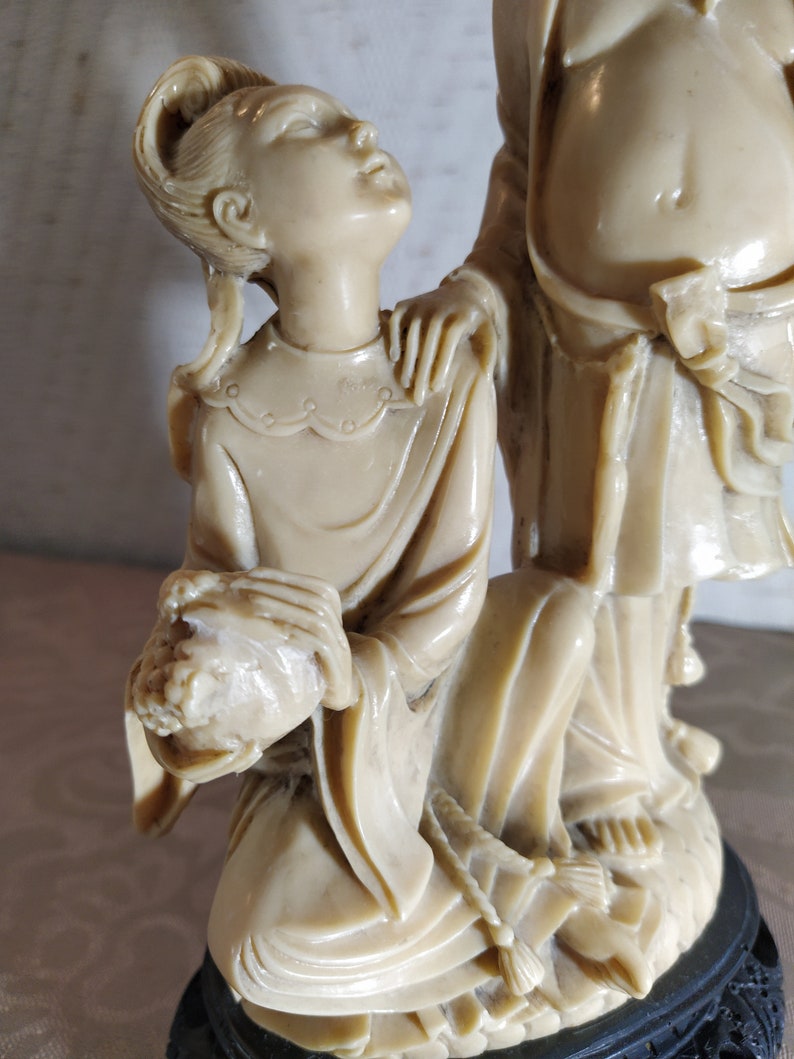 May include: A white ceramic sculpture of two figures, a man and a woman, standing on a black base. The woman is kneeling and holding a bouquet of flowers. The man is standing behind her with his hand on her shoulder.