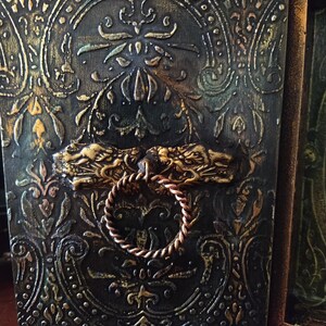 May include: A dark wooden box with a gold and green embossed design. The box has a decorative metal handle with two dragon heads and a rope-like ring.