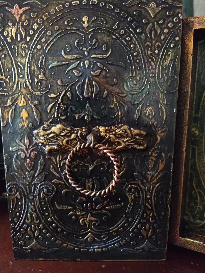 May include: A dark wooden box with a gold and green embossed design. The box has a decorative metal handle with two dragon heads and a rope-like ring.
