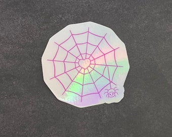 May include: A holographic sticker with a pink spiderweb design. The web has a pink heart in the center and a small pink spider at the bottom.