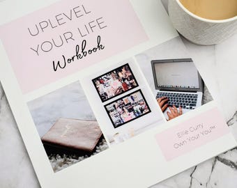 May include: A white workbook titled "Uplevel Your Life Workbook" with pink accents, featuring images of a vision board, a laptop, and a journal. A mug of coffee sits nearby. The workbook is by Ellie Curry.