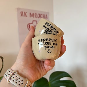Könnte beinhalten: Zwei kleine, beige Espressotassen aus Keramik, die übereinander gestapelt sind und von einer Hand gehalten werden. Jede Tasse hat einen schwarzen, handgeschriebenen Text, der "espresso makes me poop" mit einem Smiley-Gesicht lautet. Die Tassen haben kleine, bunte Blumenakzente.