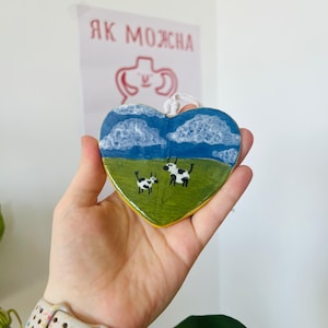 Puede incluir: Adorno de cerámica en forma de corazón con una escena pastoral. El diseño incluye un campo verde, cielo azul con nubes blancas y dos vacas. El adorno es sostenido por una mano, con una correa de reloj blanca y multicolor visible.