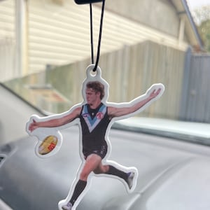 May include: A car air freshener featuring a stylized image of a football player in action, kicking a football. The player is wearing a black and teal uniform. The air freshener hangs from a black string.