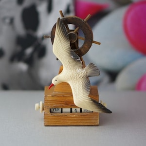Può includere: Una statuetta di gabbiano in legno con un becco rosso, posata su una base in legno con una piccola ruota in legno. La base ha una funzione di calendario con le parole "VEN" e "1" visibili.
