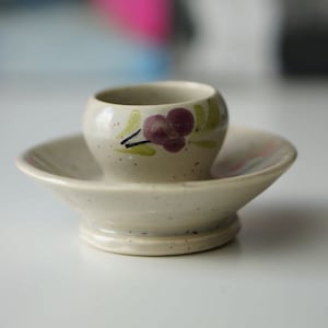 May include: A small, white ceramic egg cup with a floral design. The cup is sitting on a matching saucer.