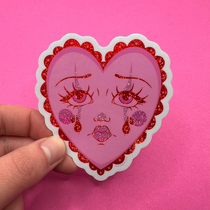 May include: A pink heart-shaped sticker with a cartoon face crying red glitter tears. The heart has a red glitter border.