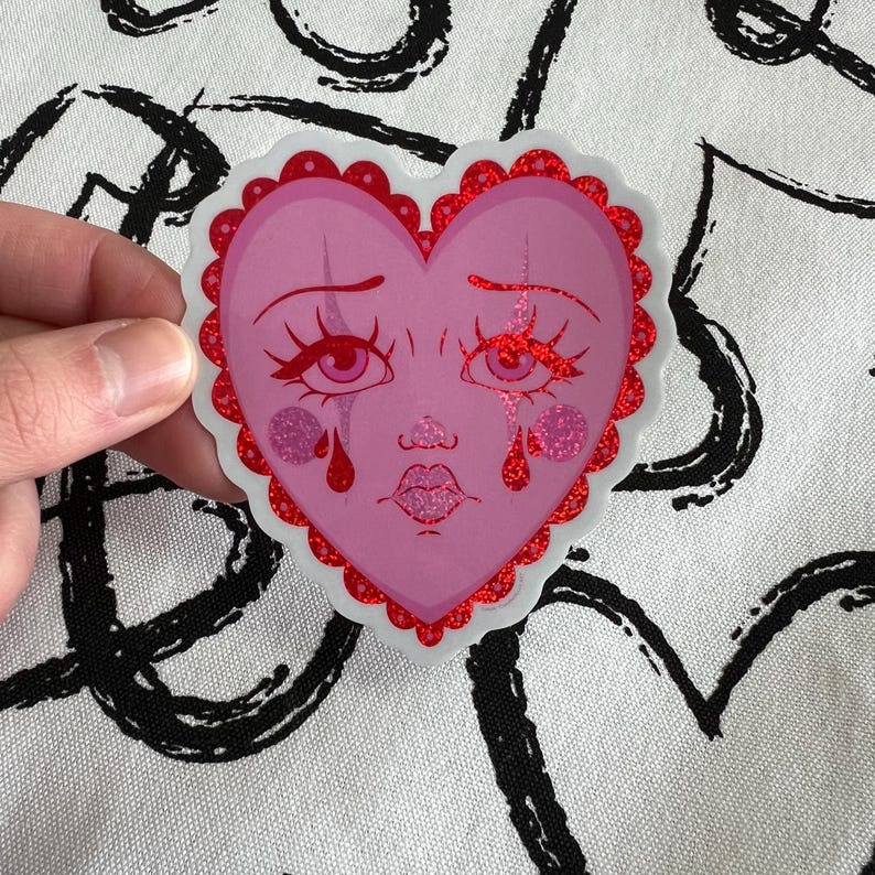 May include: A pink heart-shaped sticker with a cartoon face crying red tears. The heart has a white outline and red glitter around the edges.