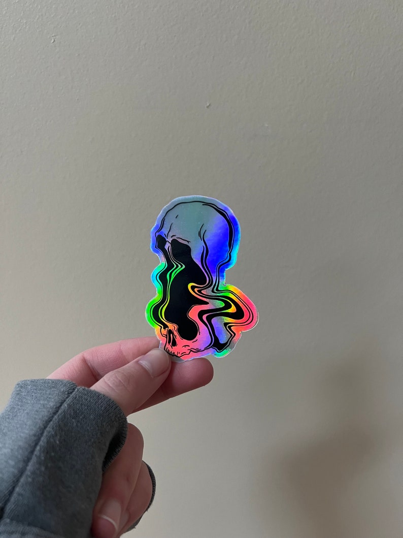 May include: A holographic sticker of a skull with a black and white design. The skull is melting and dripping with rainbow colors.