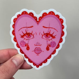 May include: A pink heart-shaped sticker with a cartoon face crying red glitter tears. The heart has a red glitter border with a scalloped edge.
