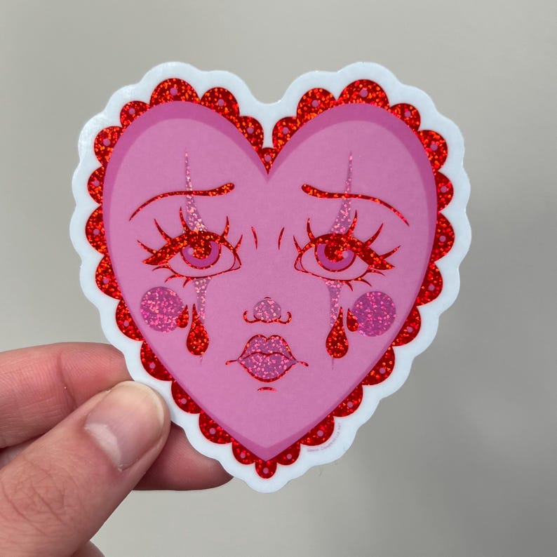May include: A pink heart-shaped sticker with a cartoon face crying red glitter tears. The heart has a red glitter border with a scalloped edge.