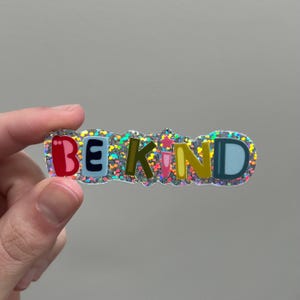 May include: A silver glitter sticker with the words "BE KIND" in colorful letters. The letters are outlined in black and have a slightly raised texture.