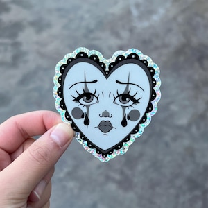 May include: A heart-shaped sticker featuring a sad clown face with teardrops. The face is light blue with black accents, including the eyes, eyebrows, and lips. The sticker has a black scalloped border and a glittery, iridescent edge.