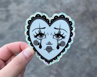 May include: A heart-shaped sticker featuring a sad clown face with teardrops. The face is light blue with black accents, including the eyes, eyebrows, and lips. The sticker has a black scalloped border and a glittery, iridescent edge.