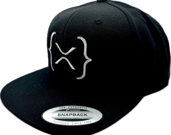May include: Black baseball cap with a white embroidered logo of curly brackets. The cap has a black brim and a black adjustable strap with a silver metal buckle. The text "THE AUTHENTIC YUPOONG SNAPBACK YUPOONG SINCE 1974" is printed on the back of the cap.