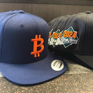 Könnte beinhalten: Zwei blaue und schwarze Baseballkappen mit einem orangefarbenen Bitcoin-Symbol und dem Text "2022 2024 Bitcoin Halving", der auf der Vorderseite gestickt ist.