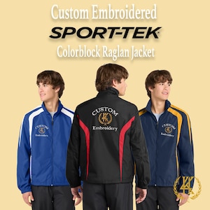 Puede incluir: Tres chaquetas raglán Sport-Tek colorblock bordadas a medida en azul, negro y marino. Las chaquetas tienen cierre de cremallera y presentan el texto "Custom Embroidery". La chaqueta negra tiene detalles en rojo.