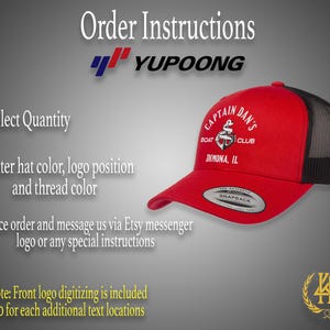 May include: Red and black trucker hat with a white embroidered logo that says "Captain Dan's Boat Club Dimona, IL". The hat has a black mesh back and a white snap closure.