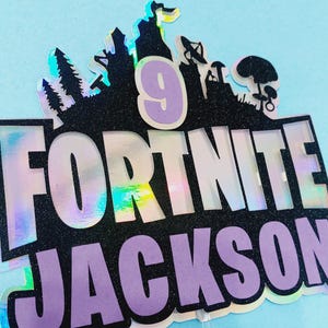 May include: A holographic cake topper with the words "Fortnite" and "Jackson" in purple and black lettering. The number "9" is in the center of the topper, surrounded by a black silhouette of a Fortnite video game scene.