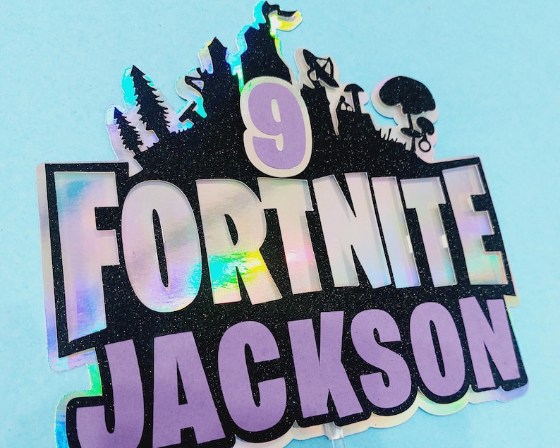 May include: A holographic cake topper with the words "Fortnite" and "Jackson" in purple and black lettering. The number "9" is in the center of the topper, surrounded by a black silhouette of a Fortnite video game scene.