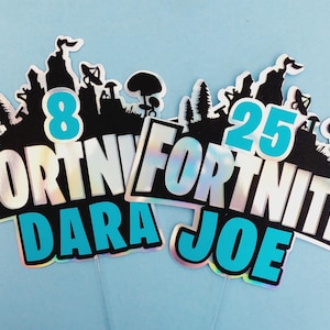 May include: Two Fortnite themed cake toppers with iridescent backgrounds. One topper says "Fortnite Dara 8" in blue and black lettering. The other topper says "Fortnite Joe 25" in blue and black lettering.