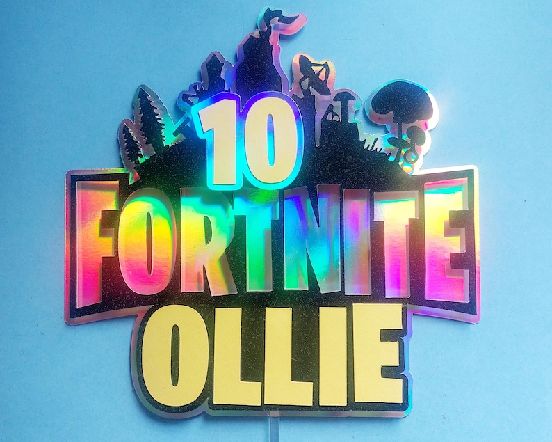 May include: A holographic cake topper with the words "10 Fortnite Ollie" in a rainbow color scheme. The topper features a silhouette of a Fortnite character and other game-related imagery.