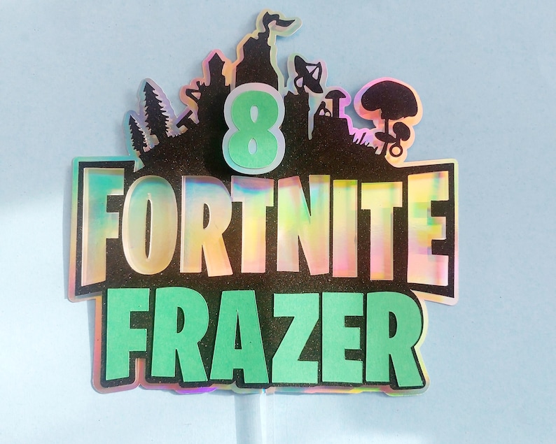 May include: A green and black cake topper with the words "Fortnite Frazer" and the number "8" in a green circle. The background is a black and iridescent design with a silhouette of a video game character.