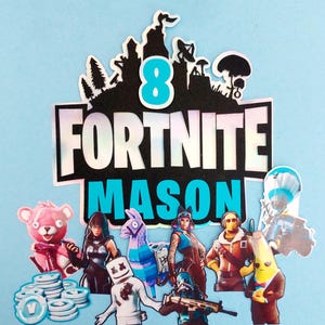 May include: A Fortnite themed cake topper with the words "Fortnite Mason 8" in blue and black lettering. The topper features various Fortnite characters, including a llama, a pink bear, and a masked figure.