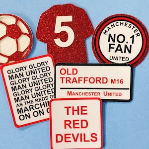 May include: A set of six red and white Manchester United themed decorations. The decorations include a football, a jersey with the number 5, a circle with the text "Manchester No. 1 Fan United", a rectangle with the text "Old Trafford M16 Manchester United", a rectangle with the text "Glory Glory Man United" and a rectangle with the text "The Red Devils".