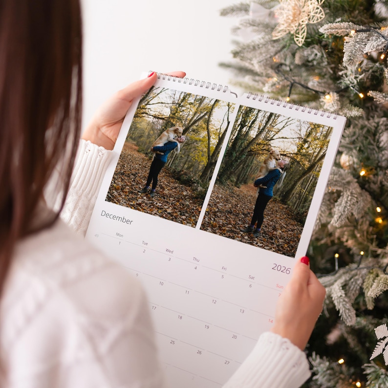 Op de afbeelding: Een persoon houdt een spiraalgebonden kalender open op december 2026. De kalender toont twee foto's van een persoon in een blauwe jas en zwarte broek in een bosrijke omgeving. De kalender is wit met zwarte tekst.