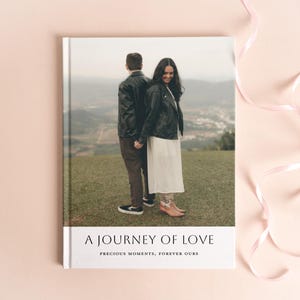 May include: A hardcover photo album with the title "A Journey of Love" and the text "Precious Moments, Forever Ours." The cover features a couple holding hands, with a scenic background. Pink ribbons are draped on the side.