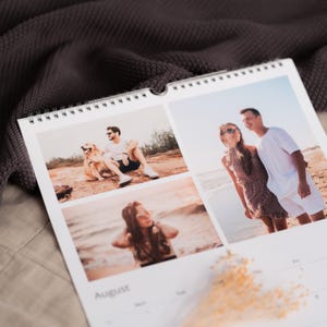 Op de afbeelding: Een spiraalgebonden wandkalender met drie foto's. De foto linksboven toont een persoon met een hond. De foto rechtsboven toont een stel op een strand. De onderste foto toont een persoon op het strand. De maand augustus staat onderaan afgedrukt.