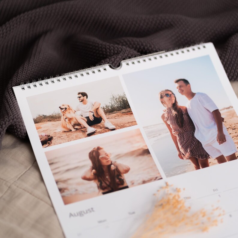 Op de afbeelding: Een spiraalgebonden wandkalender met drie foto's. De foto linksboven toont een persoon met een hond. De foto rechtsboven toont een stel op een strand. De onderste foto toont een persoon op het strand. De maand augustus staat onderaan afgedrukt.