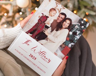 May include: A photo book with a family portrait, held by a person. The cover is white with red text that reads "The Williams Family 2025 A Year of Unforgettable Moments." The photo shows a family of three with a Christmas tree in the background.