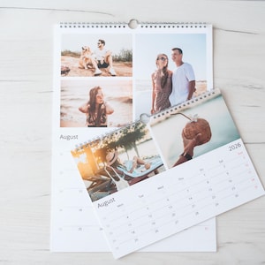 Op de afbeelding: Een spiraalgebonden kalender met foto's van mensen die van zomerse activiteiten genieten. De kalender toont de maand augustus en het jaar 2026. De kalender ligt open op een lichtgekleurd houten oppervlak.