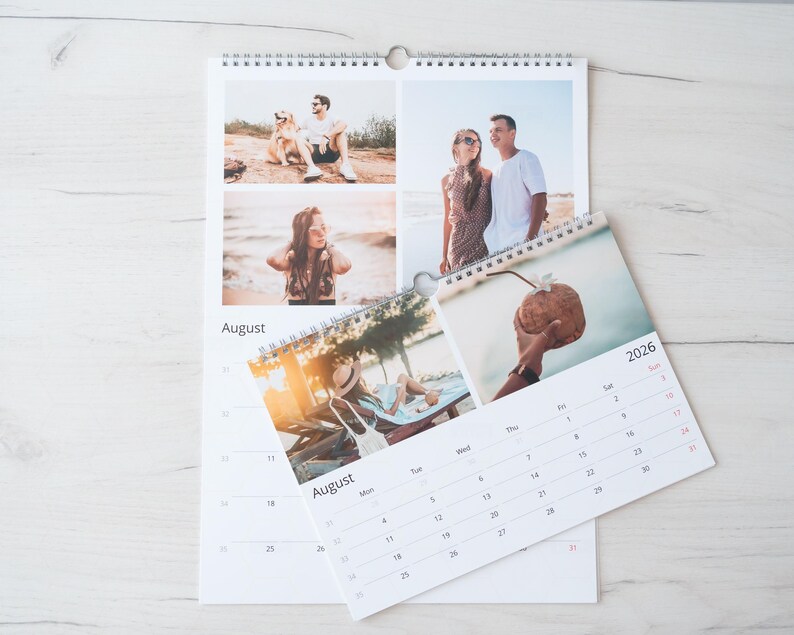 Op de afbeelding: Een spiraalgebonden kalender met foto's van mensen die van zomerse activiteiten genieten. De kalender toont de maand augustus en het jaar 2026. De kalender ligt open op een lichtgekleurd houten oppervlak.
