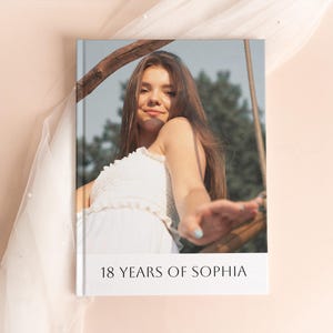 Op de afbeelding: Een hardcover fotoalbum met een foto van een jonge persoon in een witte jurk. De albumcover heeft de tekst "18 YEARS OF SOPHIA" in witte letters. De achtergrond is licht perzik van kleur.