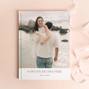 May include: A rectangular photo book with a hard cover featuring a full-bleed image of two people at the beach. The bottom white border displays the black text FOREVER BEGINS HERE and the date MAY 9, 2026 in a clean, centred serif font.