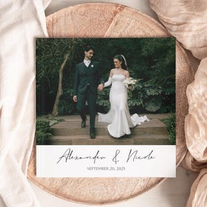 Pode incluir: Um &aacute;lbum de fotos de casamento com um casal de m&atilde;os dadas num jardim. A capa exibe os nomes "Alexander & Nicole" e a data "September 20, 2025" em escrita cursiva. O &aacute;lbum est&aacute; sobre uma bandeja decorativa.