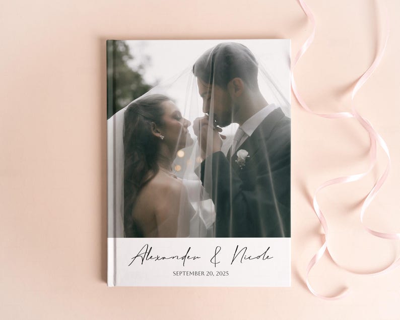 Pode incluir: Um &aacute;lbum de fotos de casamento com uma fotografia de um casal sob um v&eacute;u. A capa branca do &aacute;lbum apresenta os nomes "Alexander & Nicole" e a data "SEPTEMBER 20, 2025" em escrita elegante.