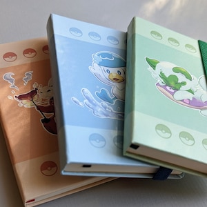 May include: Three small, colorful notebooks with cartoon characters on the covers. The notebooks are stacked on top of each other. The top notebook is green with a white and green cartoon character. The middle notebook is blue with a white cartoon character. The bottom notebook is brown with a brown cartoon character.