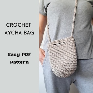 Pode incluir: Uma bolsa Aycha bege de crochê com uma alça longa. A bolsa é cilíndrica com uma pequena abertura na parte superior. O texto "CROCHET AYCHA BAG" e "Easy PDF Pattern" é exibido.