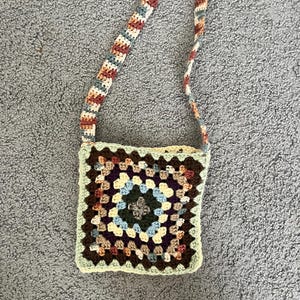 May include: A square, crocheted crossbody bag with a colorful granny square design. The bag features a strap with alternating colors of brown, blue, and cream. The bag's main body has a central square of dark green, surrounded by squares of various colors.