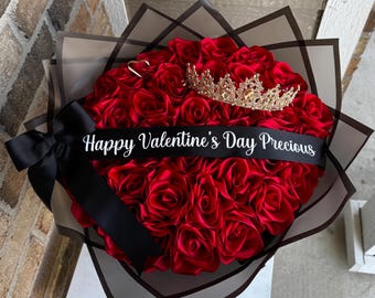 Può includere: Una composizione a forma di cuore di rose rosse, ornata da una corona d'oro e un nastro nero con la scritta "Happy Valentine's Day Precious". La composizione è avvolta in carta decorativa e legata con un fiocco nero.