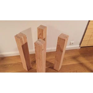 Könnte beinhalten: Vier unfertige Tischbeine aus Holz, jeweils mit einer rechteckigen Prismenform und natürlicher Holzmaserung. Die Beine stehen auf einem Holzboden und neigen sich leicht zur Mitte. Das Holz hat eine hellbraune Farbe.