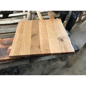 Könnte beinhalten: Eine rechteckige, unfertige Tischplatte aus Holz mit einem natürlichen Holzmaserungsmuster. Die Tischplatte besteht aus mehreren Brettern aus hellbraunem Holz, die miteinander verbunden sind. Die rechte Kante hat eine natürliche, lebende Kante. Der Tisch ruht auf einer Holzstütze.