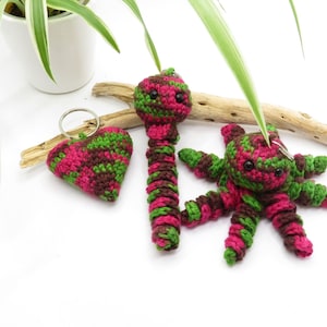 May include: Three crocheted keychains: a heart, a worm, and an octopus. The heart is pink and green, the worm is pink and green, and the octopus is pink, green, and brown.