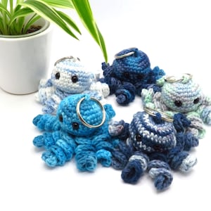 May include: Five crocheted octopus keyrings in shades of blue and white. The octopus keyrings are arranged in a circle on a white surface.