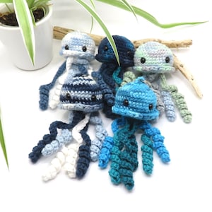 Op de afbeelding: Een verzameling handgemaakte gehaakte kwallen in verschillende tinten blauw, wit en groen. Elke kwal heeft een ronde kop met zwarte ogen en lange, vloeiende tentakels. De kwallen zijn gerangschikt op een wit oppervlak, met een plant en drijfhout op de achtergrond.